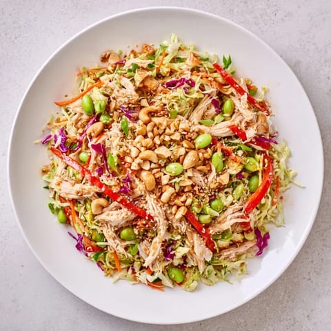 A close-up of Edamame Crunch Chicken Salad with shredded chicken, crisp cabbage, carrots, and edamame in a glossy ginger dressing, topped with toasted sesame seeds.