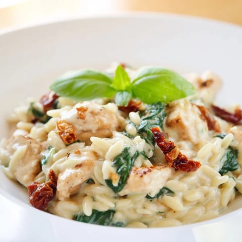 A close-up of Tuscan Chicken Orzo with creamy sauce, sun-dried tomatoes, and wilted spinach, garnished with fresh basil and Parmesan.  